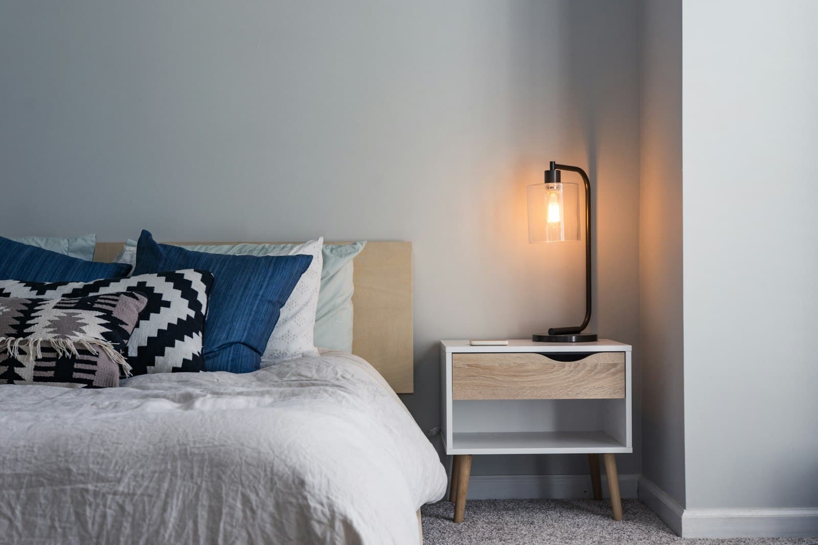 Corner of a bedroom with linen bedding, a wood headboard, and a lamp on a compact nightstand